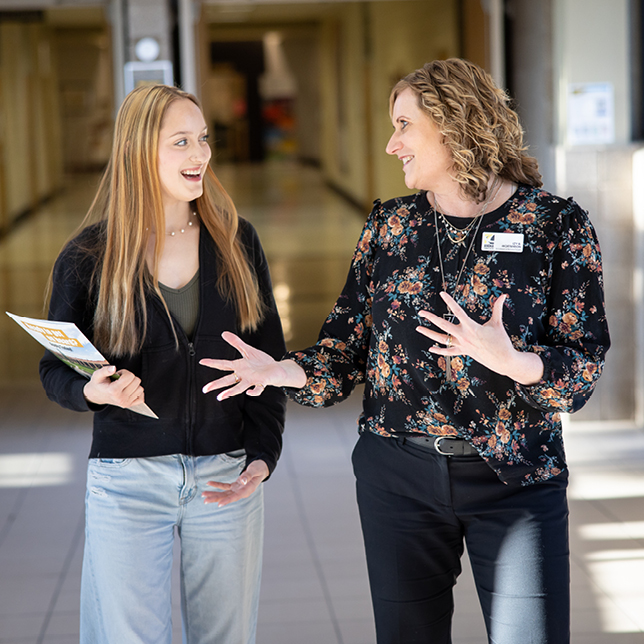 Two individuals are having a conversation in a brightly lit school hallway, one holding papers. They appear engaged and happy, standing near classrooms.