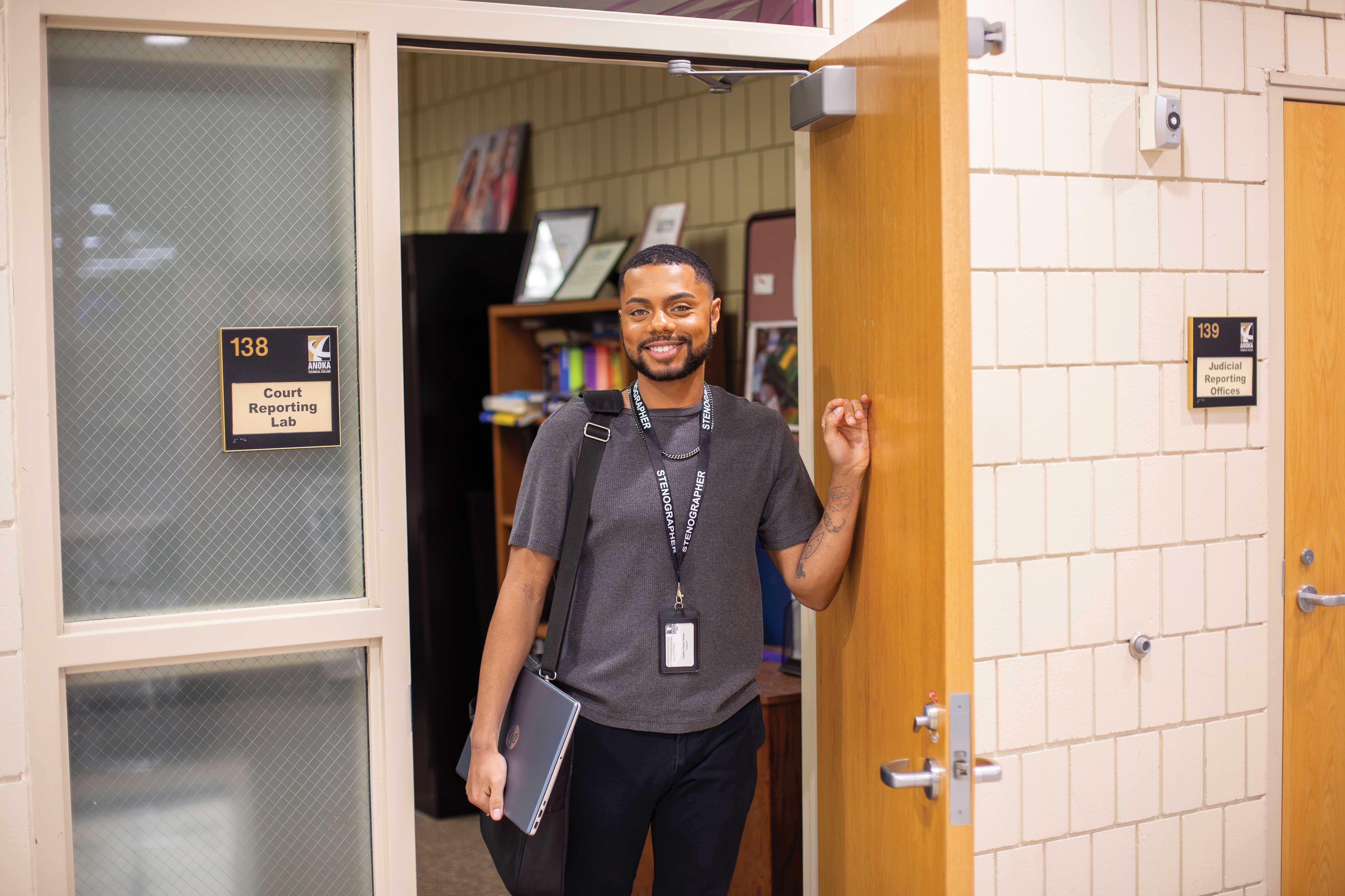 Person with a lanyard and tablet standing at the open door of a school classroom labeled 'Court Reporting Lab.'