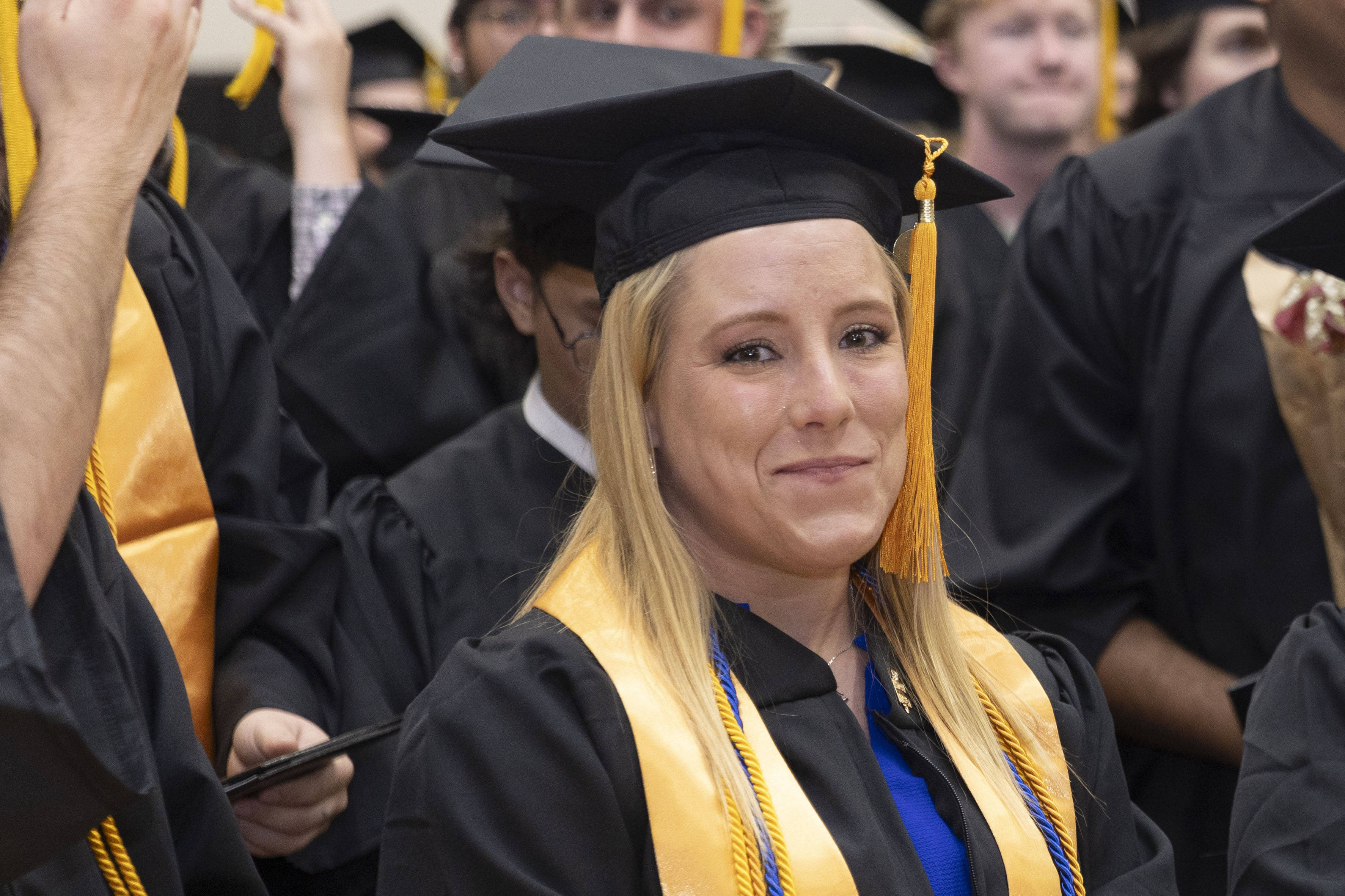 Graduate wearing a black cap and gown with a yellow honor cord smiles during a commencement ceremony.